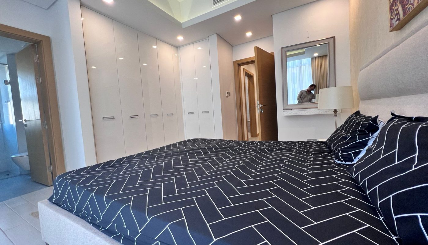 Modern bedroom with a geometric-patterned bedspread, white built-in wardrobes, a wall mirror, and an open door leading to a bathroom.