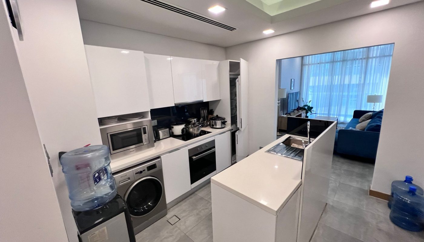 Modern kitchen with white cabinets, appliances including a washing machine, oven, and microwave, and an island; open doorway leads to a living room with blue sofa and large windows.