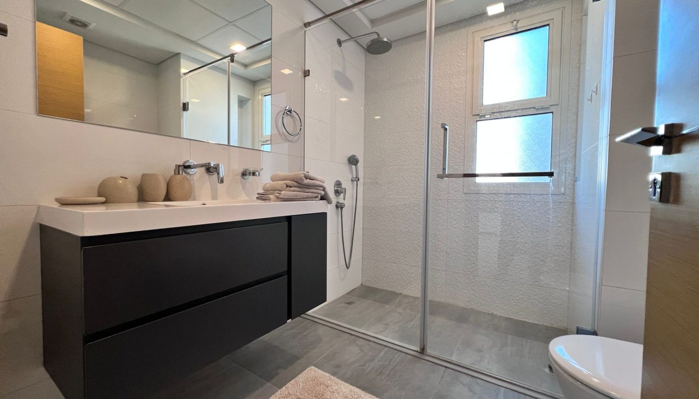 Modern bathroom with a large mirror, double sink, black vanity, walk-in glass shower, stacked towels, window, and gray tile floor.