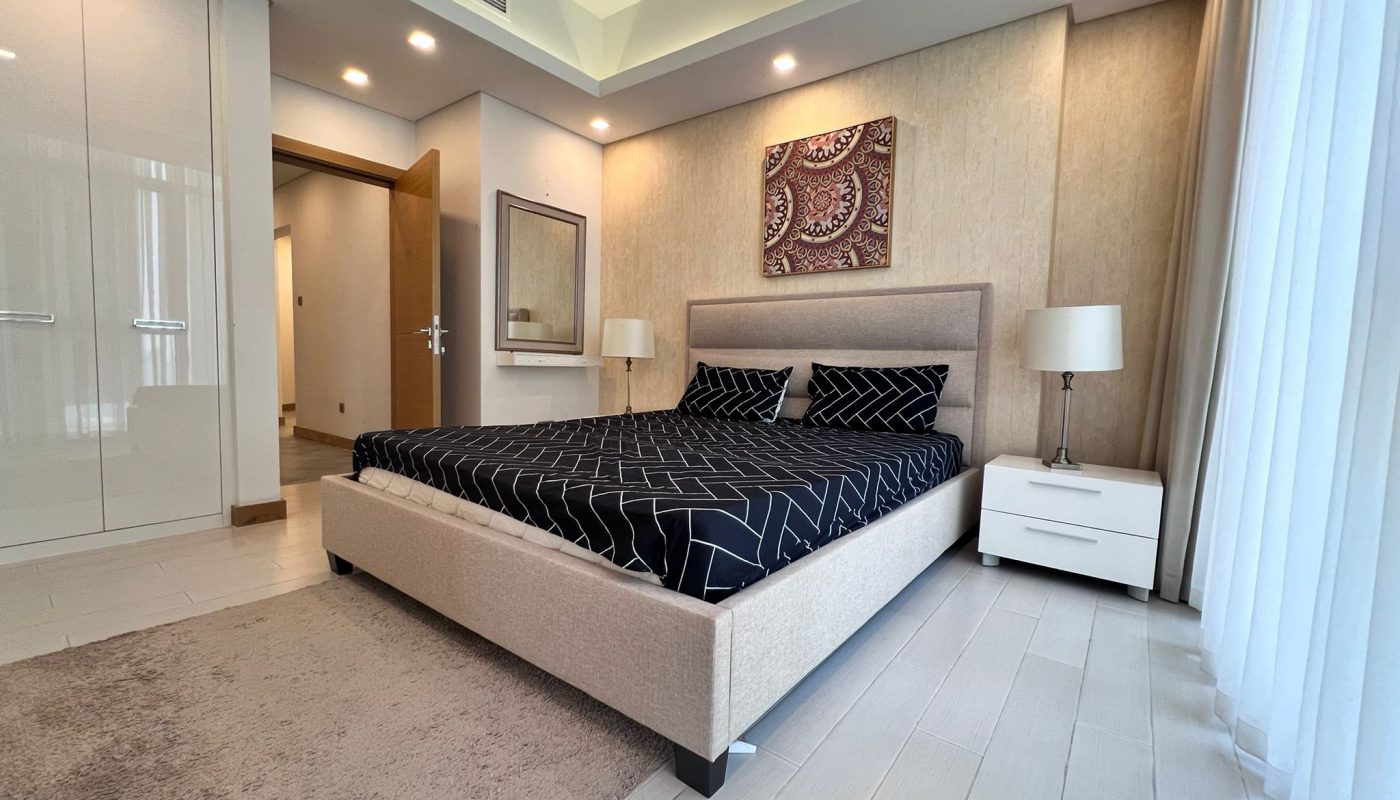 Modern bedroom with a beige upholstered bed, black geometric bedding, two white nightstands with lamps, a wall mirror, and a decorative artwork above the bed.