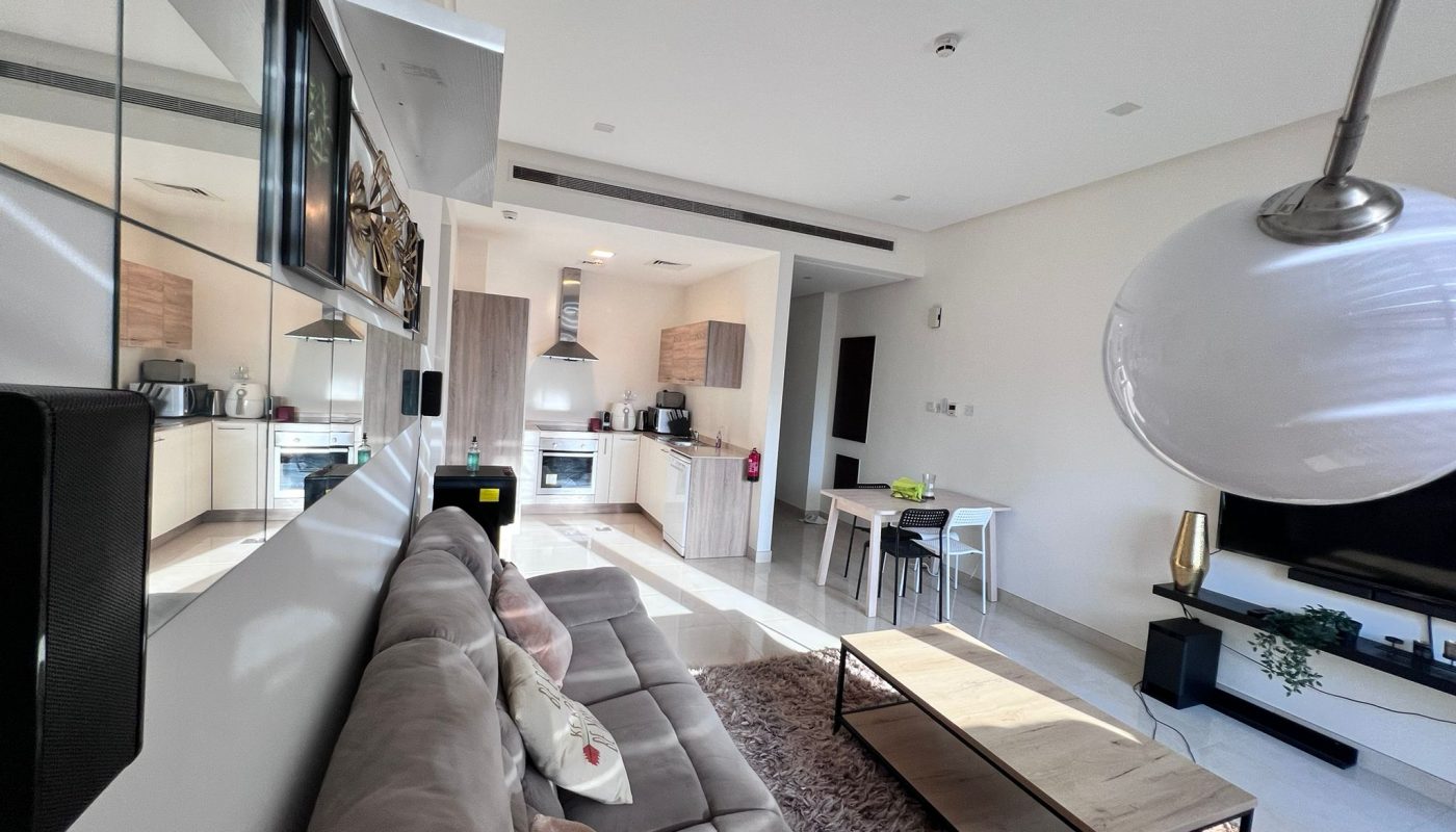 A modern, open-plan living room and kitchen with a grey sofa, coffee table, dining table, wall-mounted TV, and kitchen appliances. Sunlight streams in from the left.
