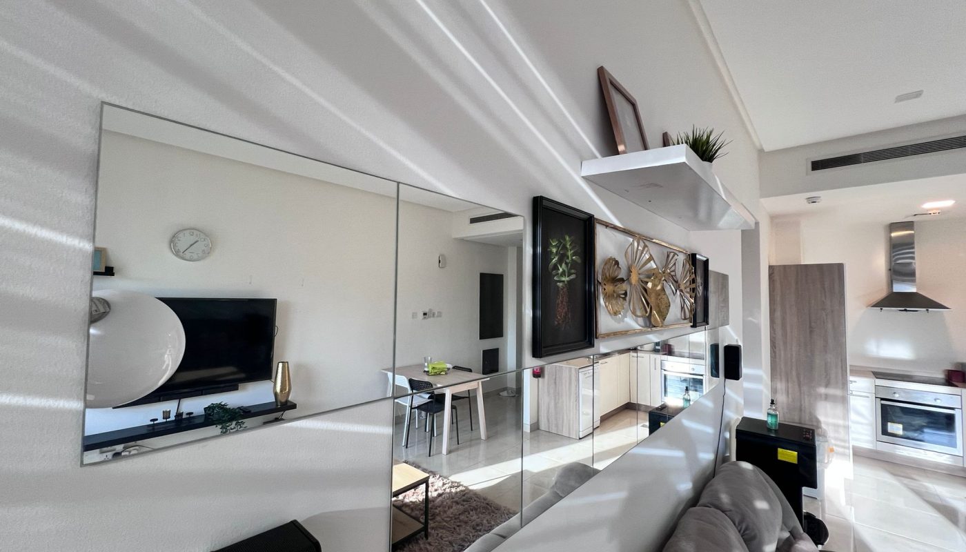 Modern living room with mirrors on the wall reflecting the kitchen and dining area, featuring a gray sofa, shelves with decor, and natural light streaming in.