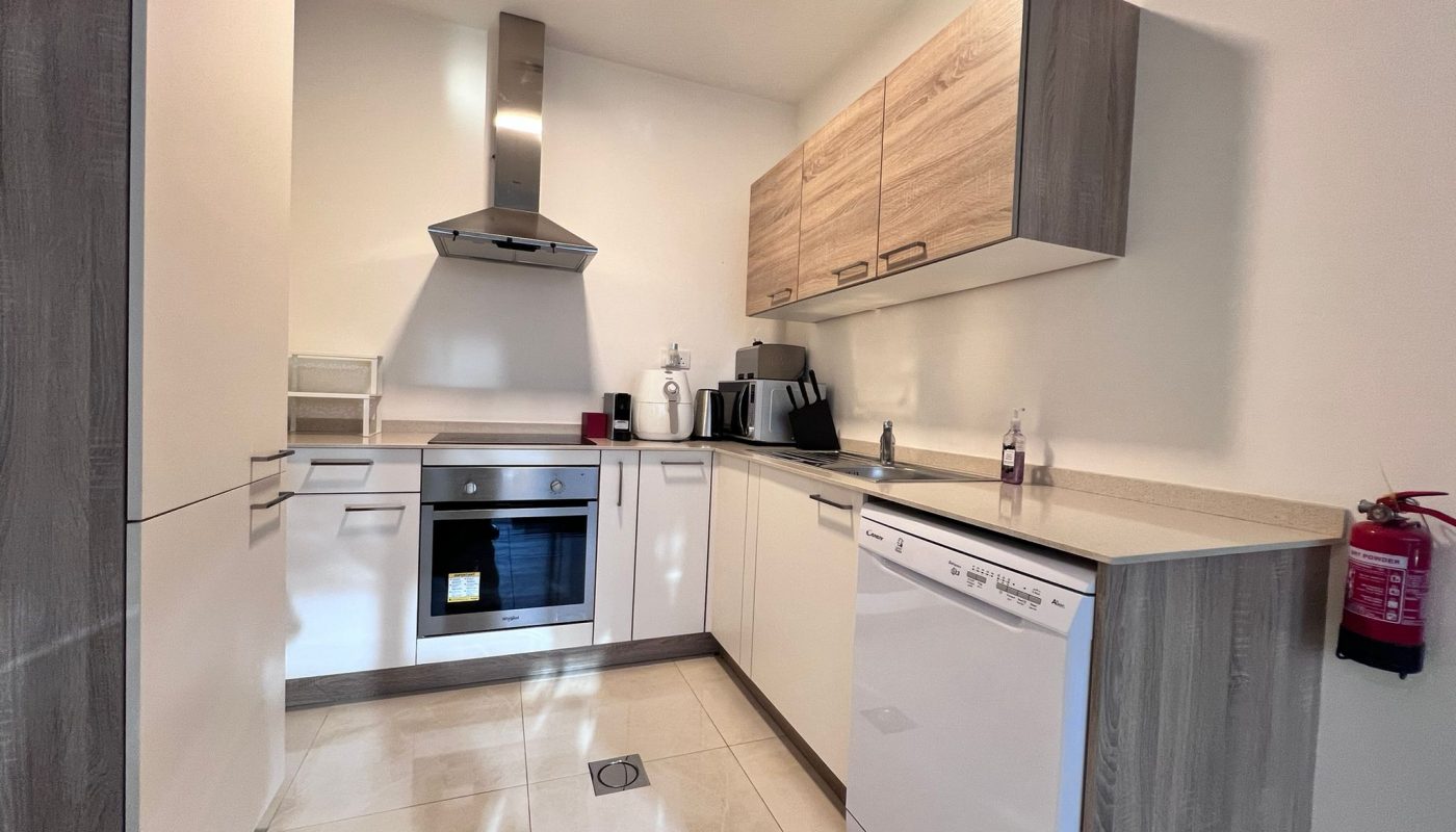 Modern kitchen with light wood cabinets, white appliances, an oven, stovetop, dishwasher, and countertop appliances. A fire extinguisher is mounted on the wall.