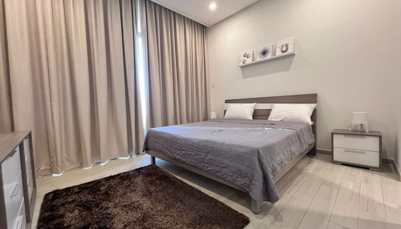 A modern bedroom with a double bed, grey bedding, brown rug, white nightstands, wall-mounted shelves, light wood flooring, and beige curtains covering a tall window.