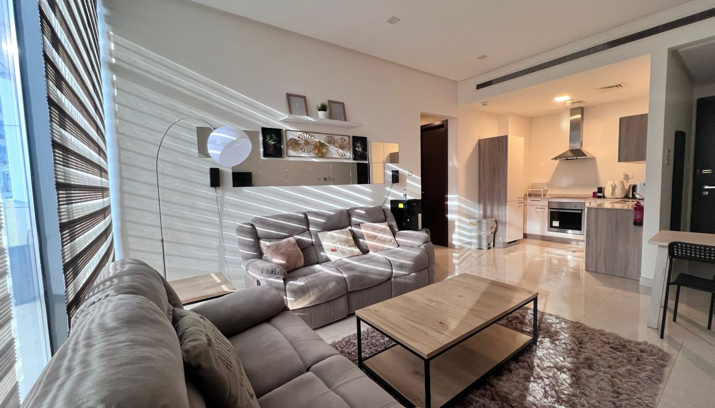Modern living room with two gray sofas, a wooden coffee table, a floor lamp, and an open kitchen in the background. Sunlight streams through large window blinds, casting shadows.