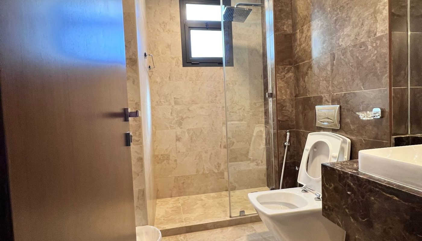 Modern bathroom with brown marble walls and floor, glass shower enclosure, toilet with open lid, bidet, sink, and a waste bin near the entrance.