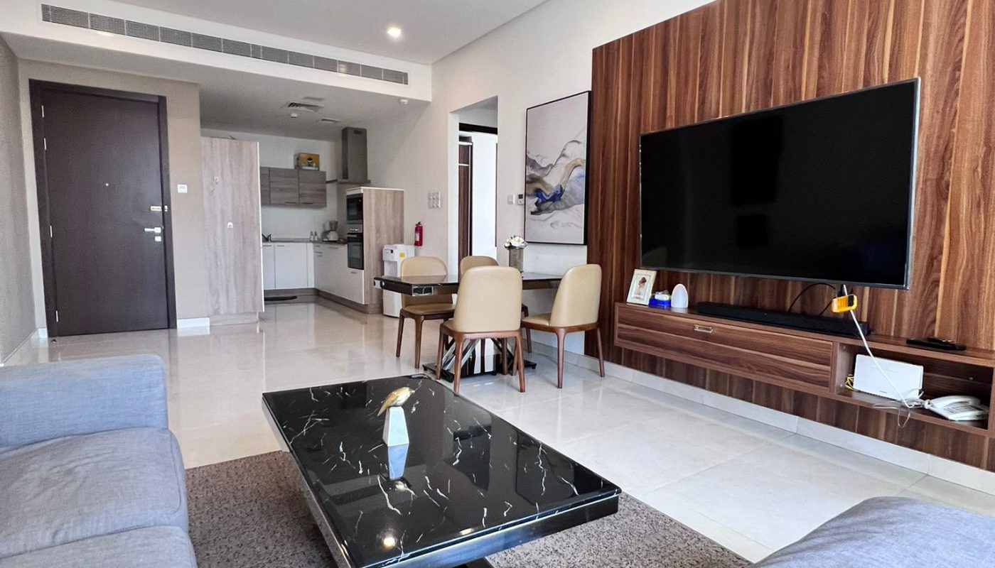 Modern living room with grey sofas, a black marble coffee table, wall-mounted TV on wood panel, and a small dining area near an open kitchen in the background.
