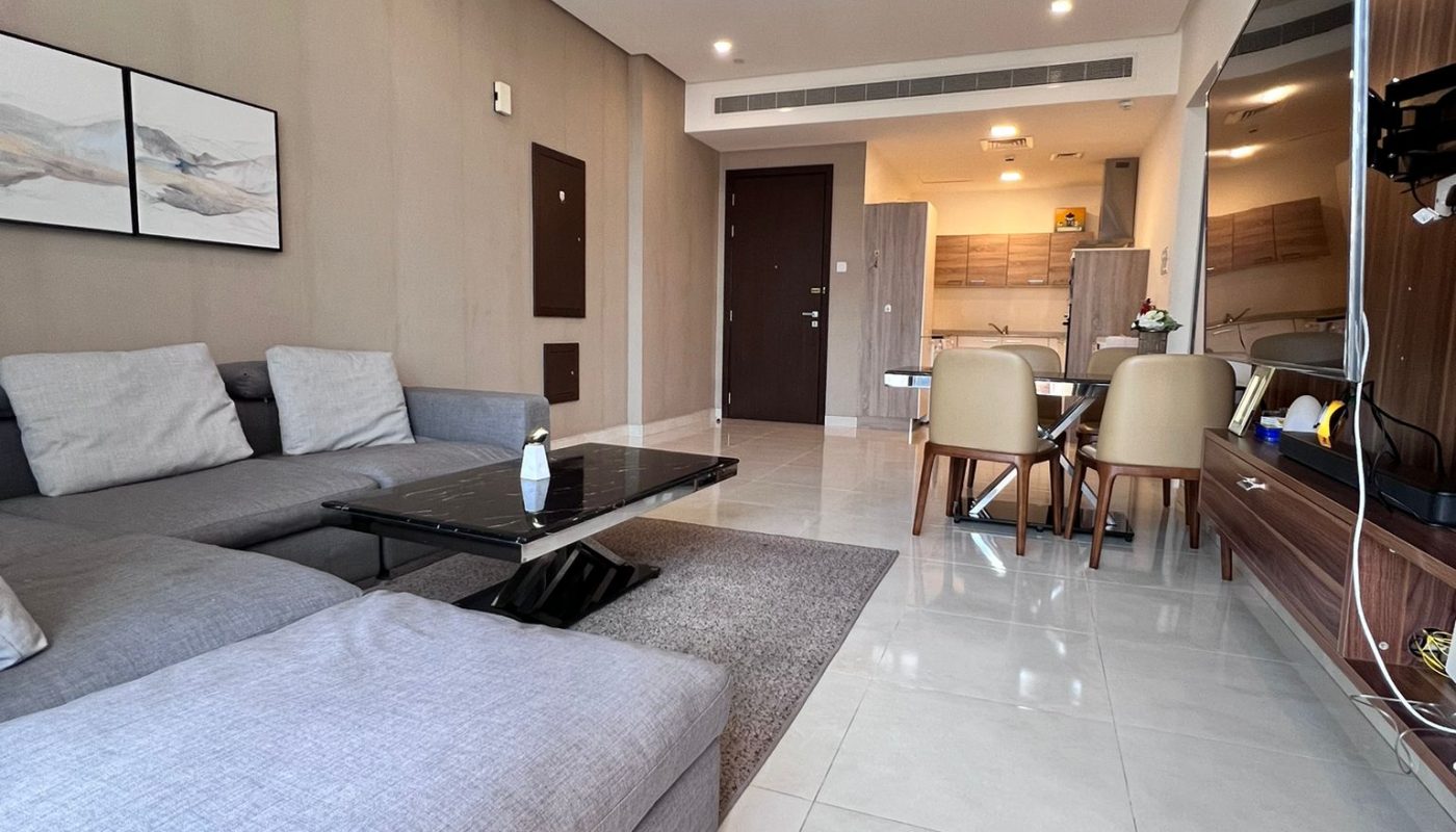 Modern living room with a gray sectional sofa, glass coffee table, dining area, and kitchenette in the background. Neutral tones and minimal decor.