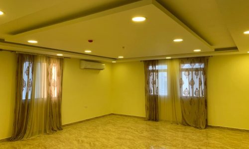 An empty room with beige walls, marble flooring, recessed lighting, two windows with sheer brown curtains, and an air conditioning unit mounted on the wall.