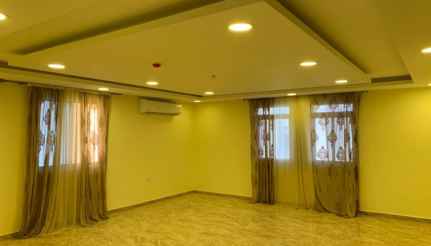 An empty room with beige walls, marble flooring, recessed lighting, two windows with sheer brown curtains, and an air conditioning unit mounted on the wall.