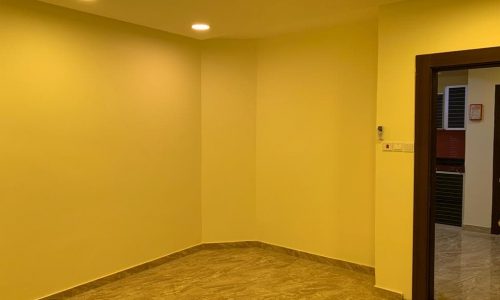 A small room with beige walls and marble flooring, illuminated by ceiling lights. There is a doorway leading to another space.