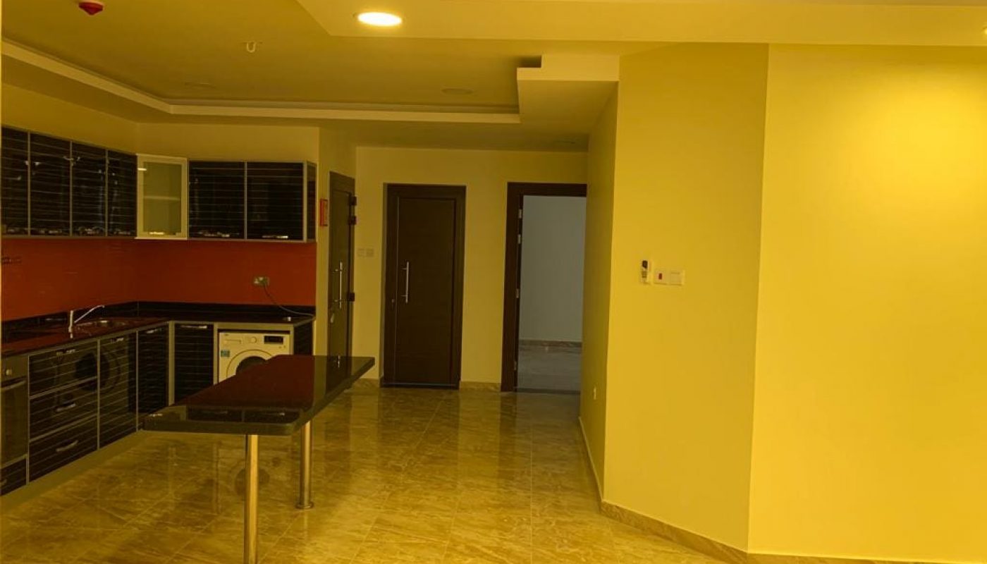 An empty kitchen and dining area with tiled floors, beige walls, dark wooden cabinets, and a black countertop. An open doorway leads to another room.