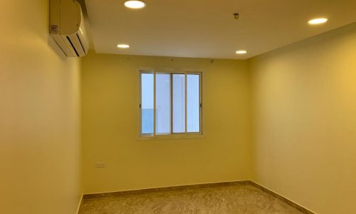 A small, empty room with beige walls, tiled floor, a window, ceiling lights, and an air conditioner mounted on the wall.