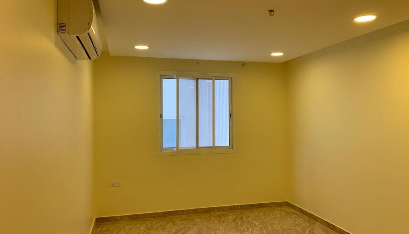 A small, empty room with beige walls, tiled floor, a window, ceiling lights, and an air conditioner mounted on the wall.