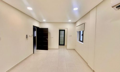 A bright, empty room in the Shakurah Spacious Triplex Apartment with white tiled floors and recessed ceiling lights. The room features a window with a blind, an air conditioning unit, and two doors: one black and partially open, one white and closed.