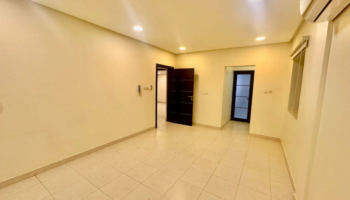 An empty room with tiled flooring, cream-colored walls, recessed ceiling lights, a doorway on the left, and another doorway on the far wall. An air conditioning unit is mounted on the right wall. This spacious Triplex Apartment in Shakurah offers ample potential for customization and comfort.