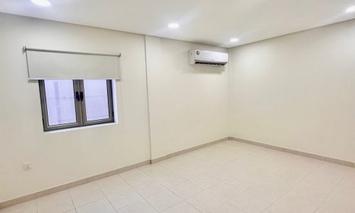 A clean, spacious room with white walls, tiled floor, a window with a roller blind, an air conditioning unit on the wall, and recessed ceiling lights in the Shakurah Triplex Apartment.