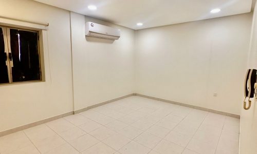 An empty room with white tiled flooring, white walls, a window with vertical blinds, an air conditioner mounted on the wall, and ceiling lights turned on. This spacious area in the Shakurah Triplex Apartment is both modern and inviting.