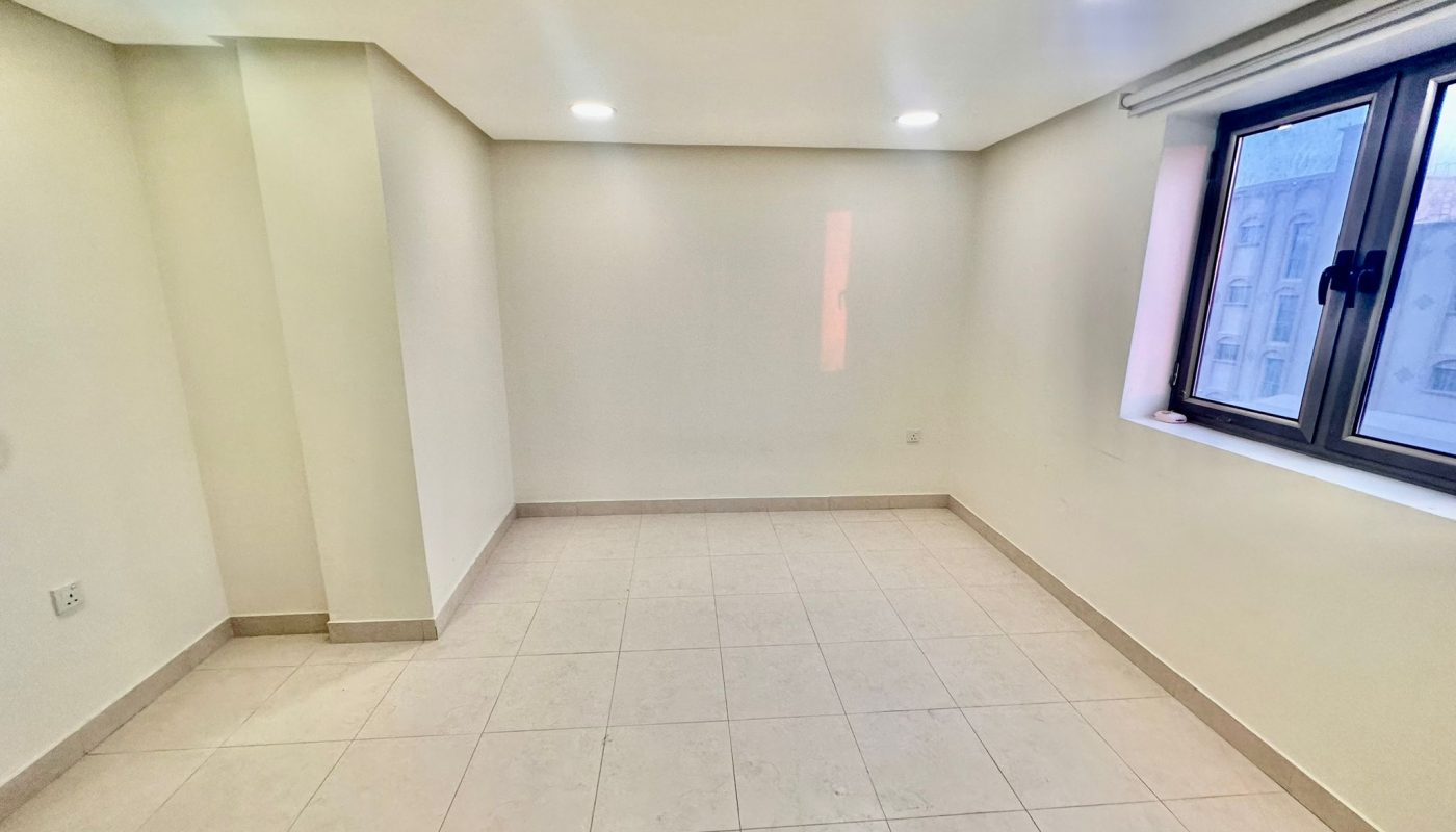 An empty, small room in a Shakurah triplex apartment features tiled floors, white walls, a window on the right letting in some natural light, and ceiling lights turned on.