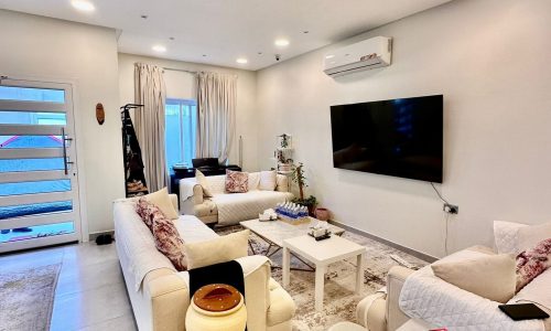 A modern living room featuring cream sofas with red cushions, a large wall-mounted TV, an air conditioner, and natural light from a glass door and windows. The space is enhanced by various furnishings and decorations, creating a cozy yet stylish ambiance.