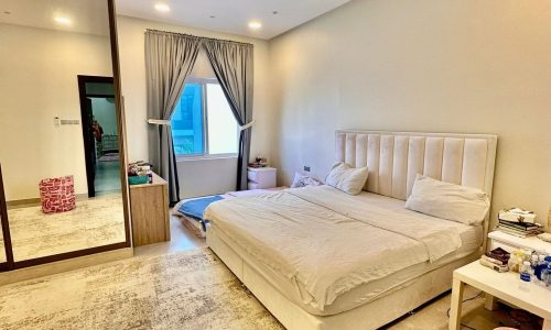 A bedroom with a large bed, beige headboard, side tables, a mirrored wardrobe, and a window with grey curtains. Various items are scattered on the floor and tables, giving the space an auto draft feel.