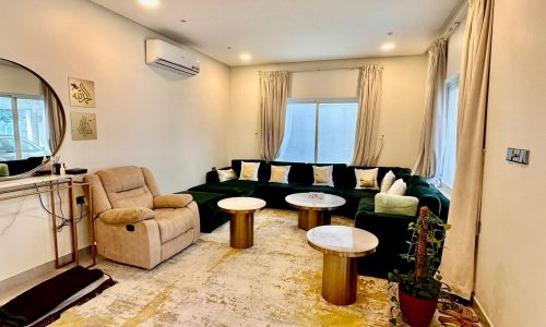 A cozy living room in a private villa in the Saar Area features a green L-shaped sofa, recliner chair, three round coffee tables, wall-mounted air conditioner, mirror, and large windows with cream curtains.