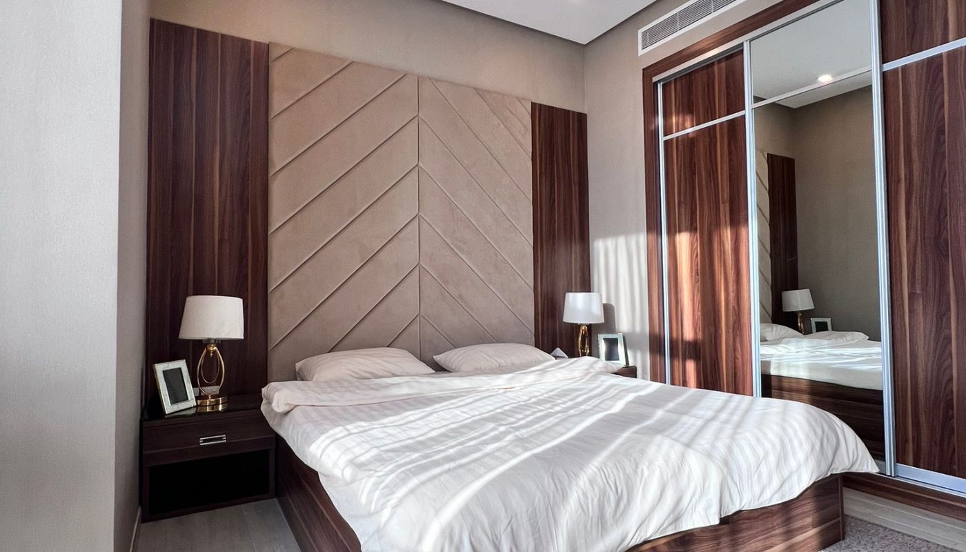 Modern bedroom with a wooden bed frame, white bedding, two nightstands with lamps, and a mirrored wardrobe reflecting sunlight and floor.