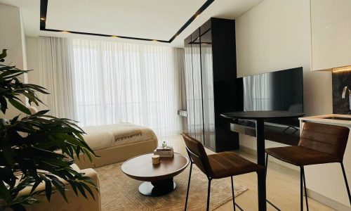 A modern living room with a round table, two chairs, a sofa, a TV, and large windows covered by sheer curtains, letting in natural light. A potted plant is placed on the left.