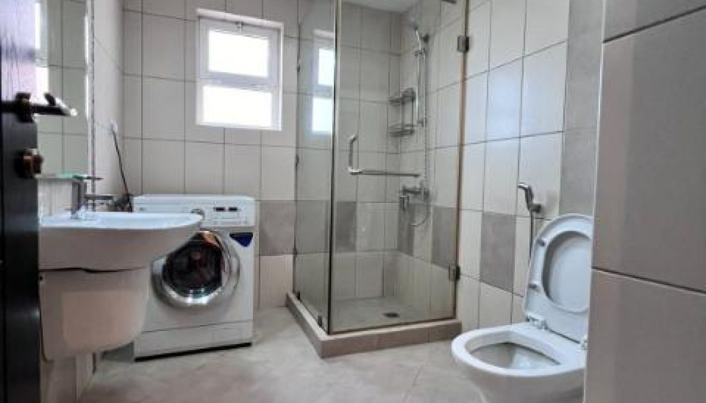 A modern bathroom with a glass shower, toilet, bidet, wall-mounted sink, and a washing machine under two small windows.