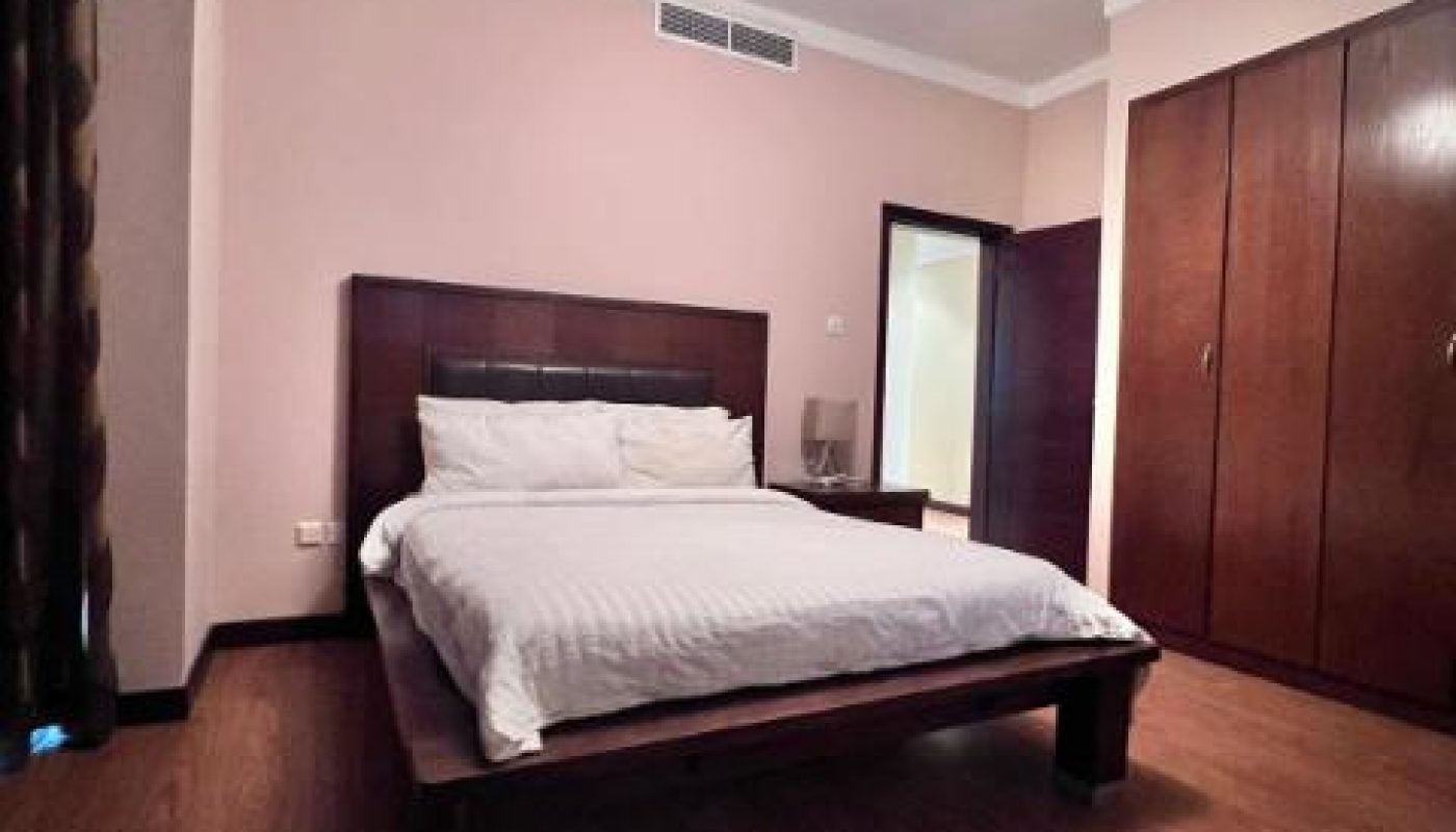 A bedroom with a wooden floor, a double bed with white bedding, built-in wooden wardrobes, and a ceiling light fixture.