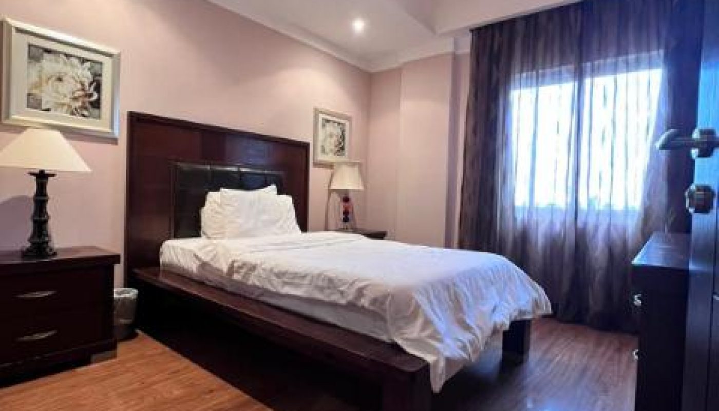 A neatly made single bed in a bedroom with wooden furniture, framed art on the walls, a lamp on each nightstand, and a window with sheer dark curtains.
