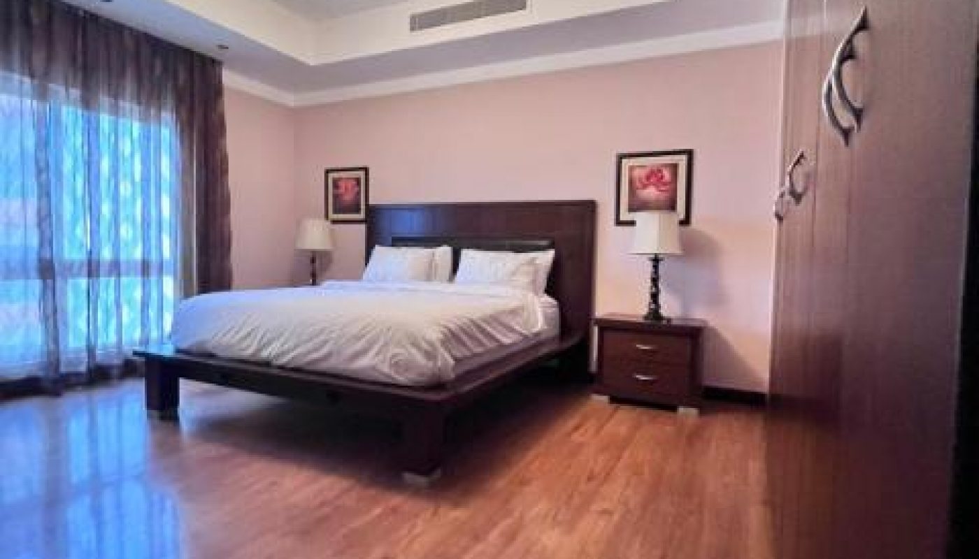 A bedroom with a large bed, two bedside tables with lamps, wooden flooring, large wardrobe, and a window with sheer curtains.