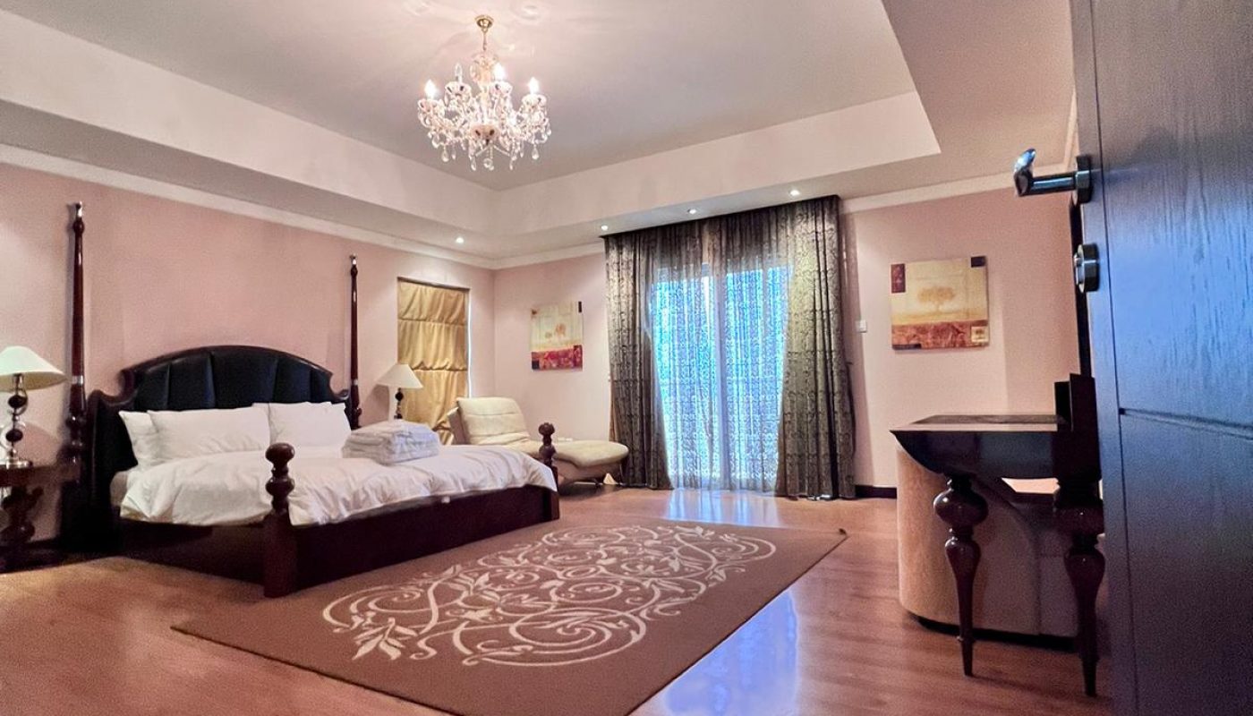 Spacious bedroom with a large bed, chandelier, wooden floor, area rug, desk, and floor-to-ceiling curtained windows letting in natural light.
