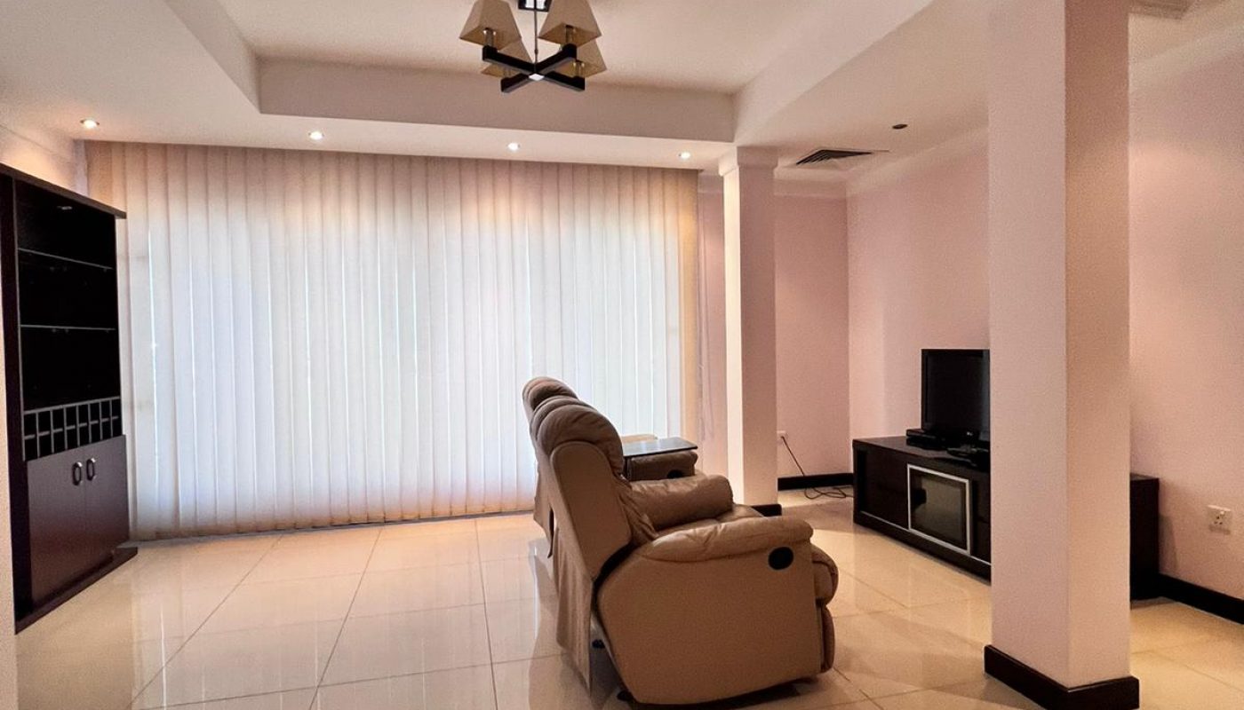 Spacious living room with tiled floor, two beige recliner chairs facing a TV on a stand, large vertical blinds covering windows, and minimal furnishings.