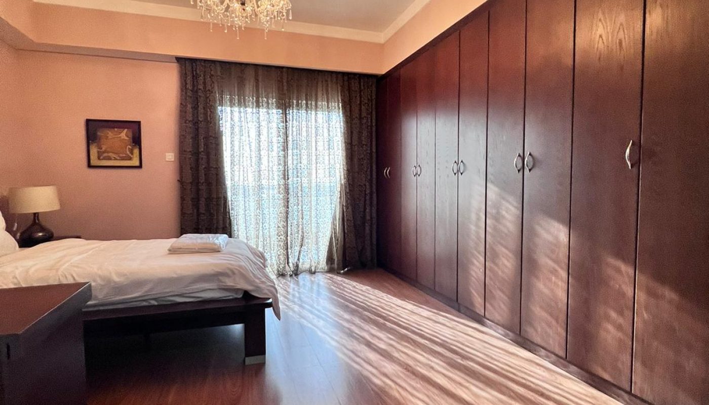 A bedroom with wood flooring, a double bed, a large wardrobe, a bedside table with a lamp, floor-length curtains, and a chandelier hanging from the ceiling.
