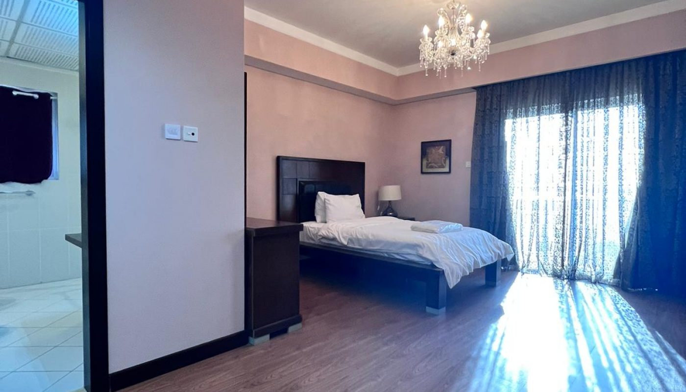 A spacious bedroom with wooden flooring, a double bed, nightstands, a chandelier, and sunlight streaming through glass doors covered by dark curtains.