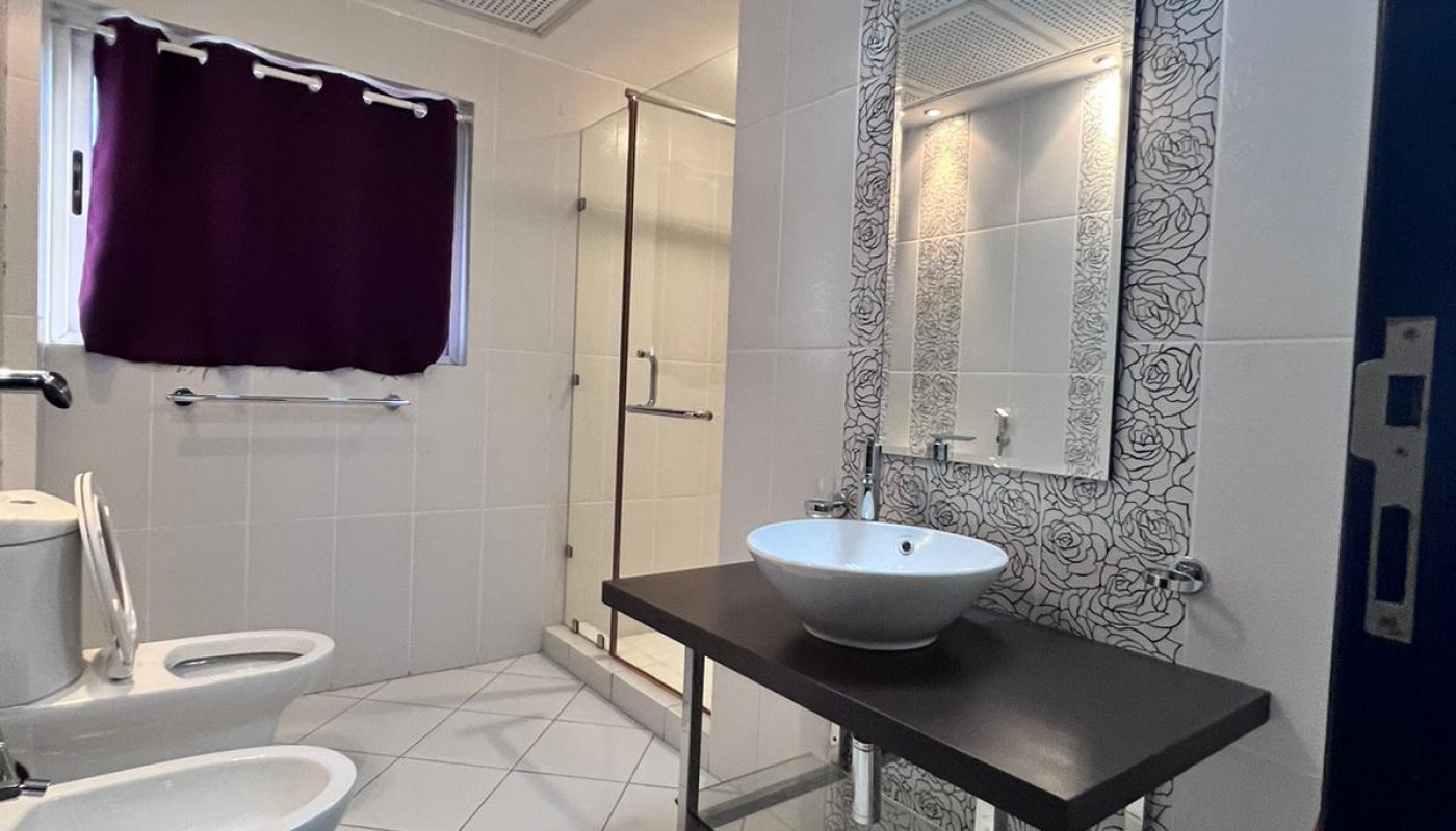 A modern bathroom with a bidet, toilet, glass shower, and a sink on a dark countertop below a lit mirror; purple curtains cover the window.