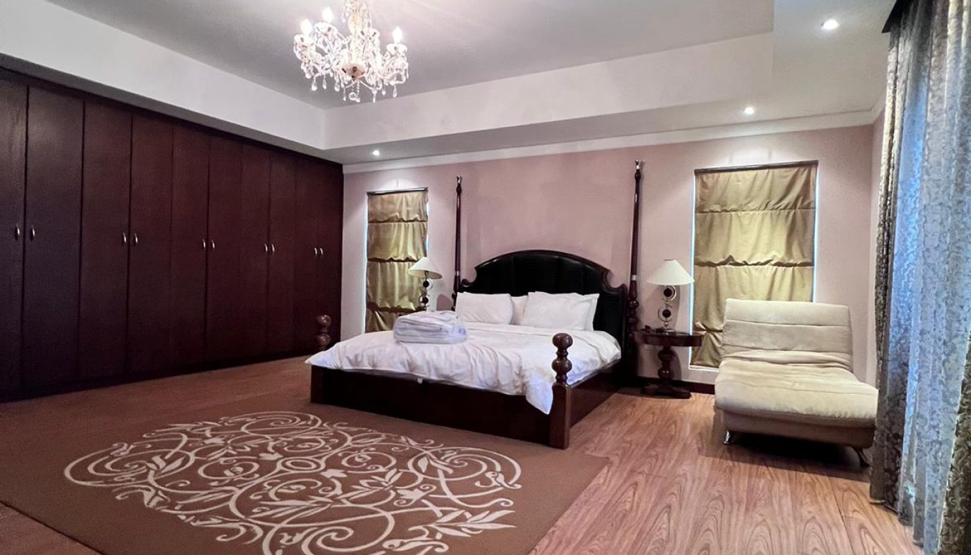 Spacious bedroom with a large wooden bed, matching side tables, a white chaise lounge, wall-to-wall wardrobes, a brown rug, and a chandelier on the ceiling.