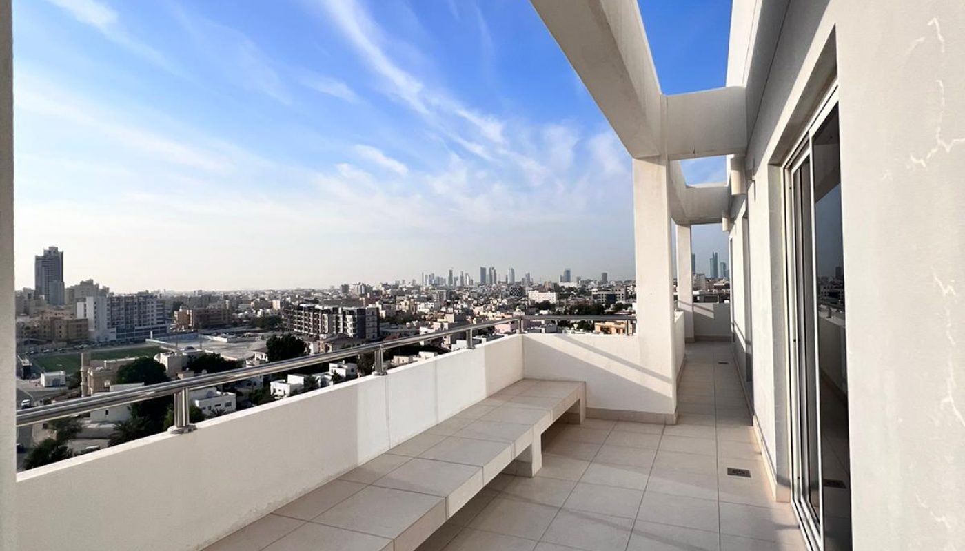 Spacious rooftop terrace with built-in white benches, tiled floor, and city skyline visible in the distance under a partly cloudy sky.