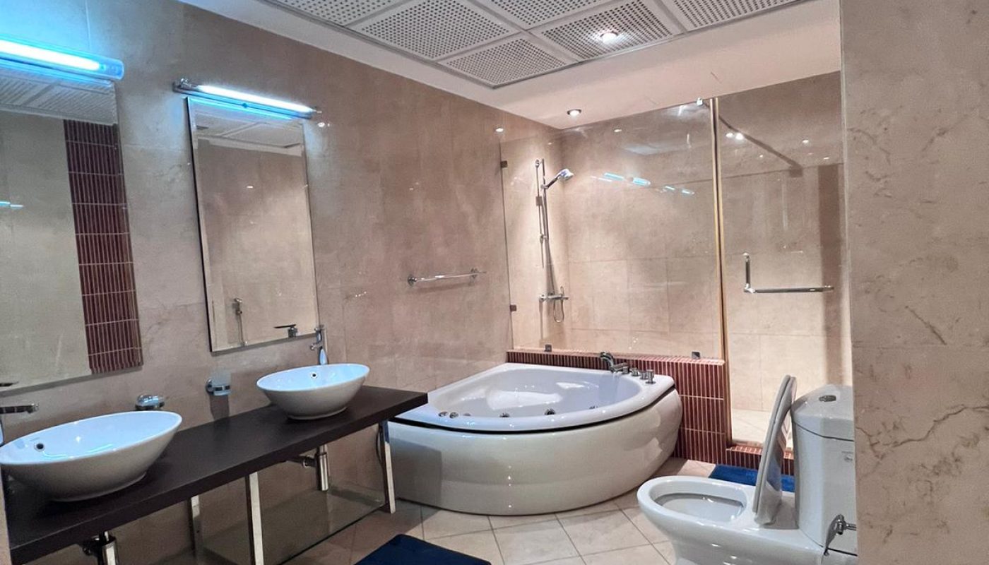 Modern bathroom with two sinks, a round bathtub, glass-enclosed shower, toilet, and bidet on tiled floor. Large mirrors and beige walls.
