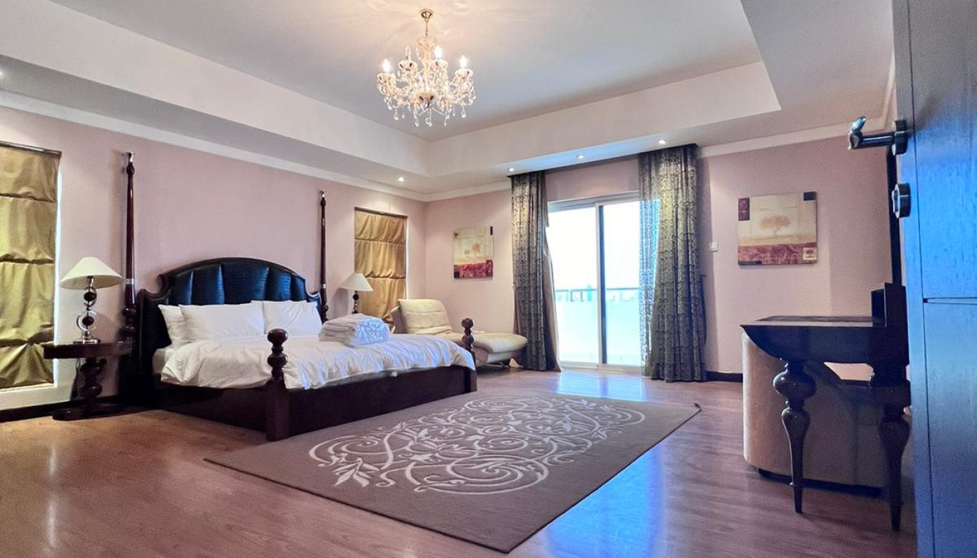 Spacious bedroom with a large bed, wooden floor, chandelier, desk, armchair, rug, and glass doors leading to a balcony with curtains.