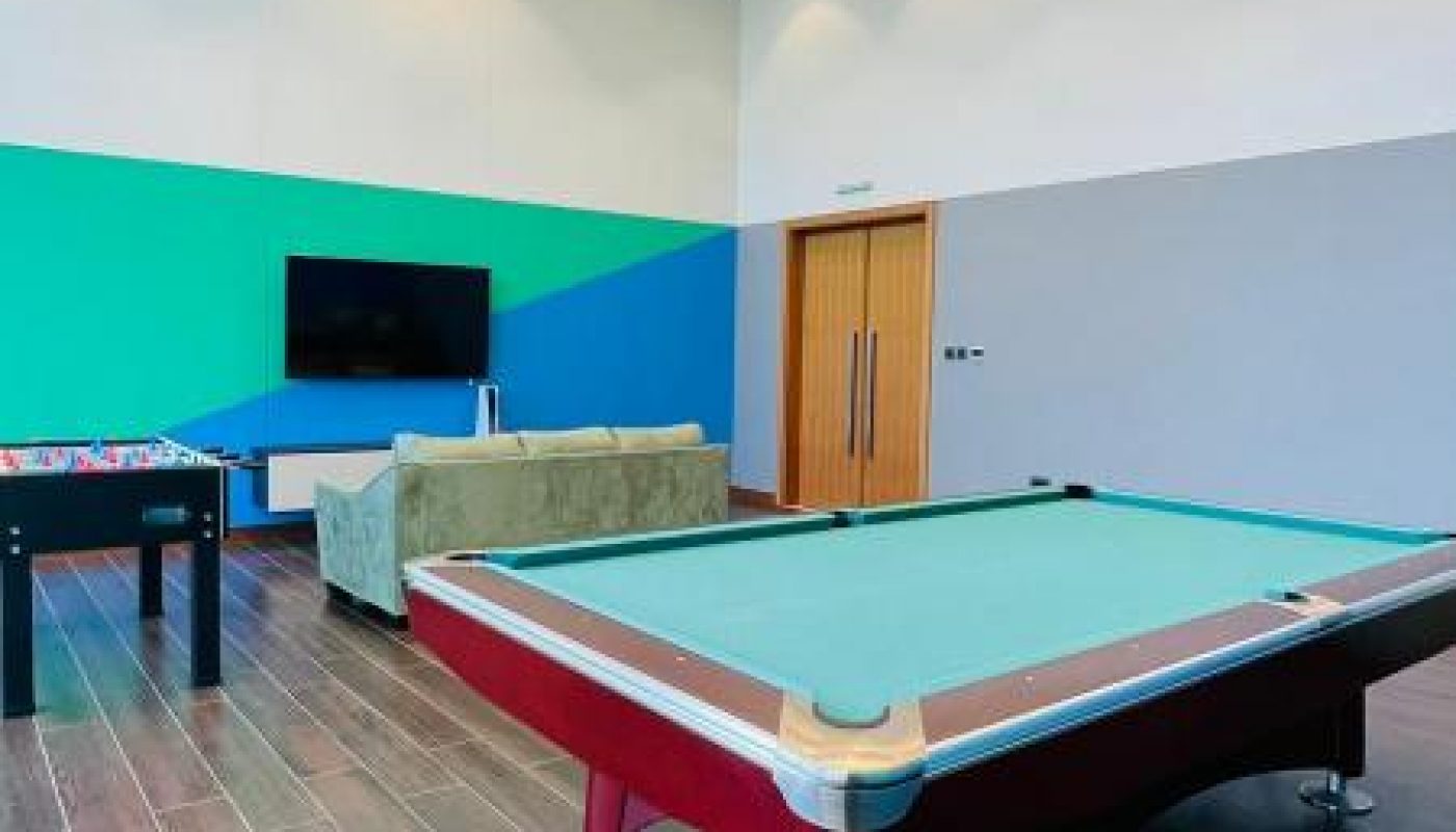 A modern game room with a pool table, foosball table, sofa, and wall-mounted TV on wood-patterned flooring.
