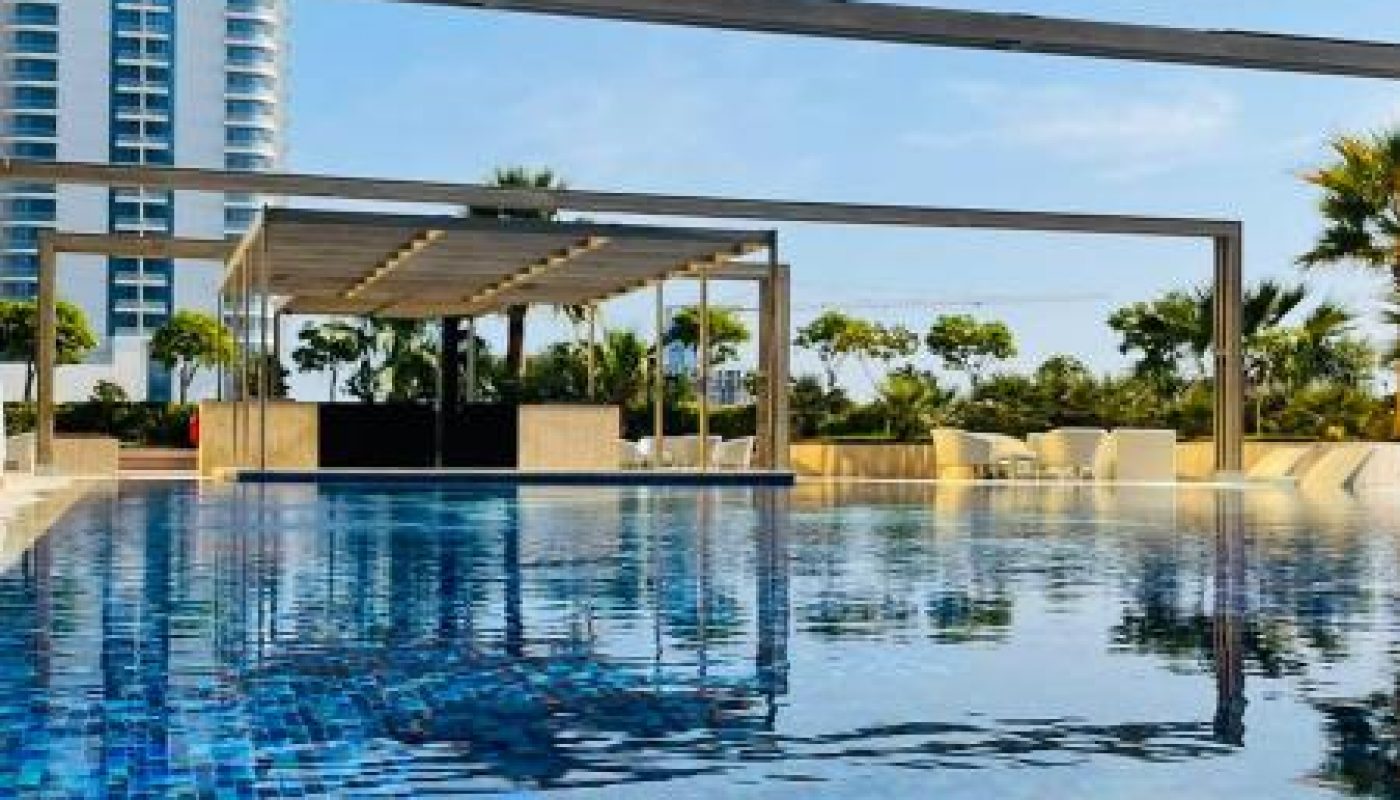 A clear blue swimming pool in the foreground, with a modern high-rise building and trees in the background under a blue sky.