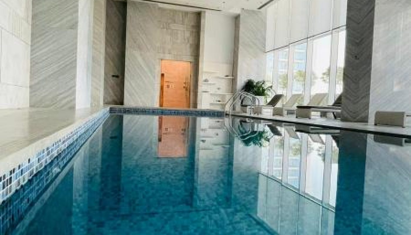 Indoor swimming pool with blue tiles, lounge chairs, and large windows letting in natural light. Marble walls and a high ceiling frame the serene space.