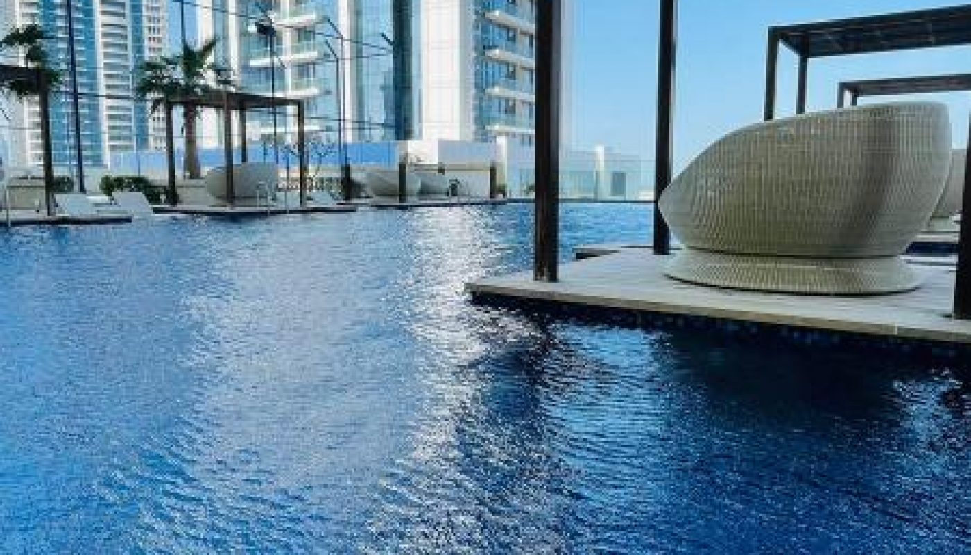 A modern high-rise building stands behind a blue swimming pool with covered lounge areas and wicker seating on a sunny day.