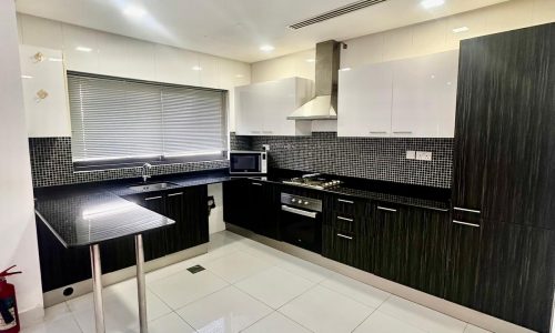 Modern kitchen with white tile flooring, black countertops, white cabinets, and stainless steel appliances. A small breakfast bar is in the foreground.