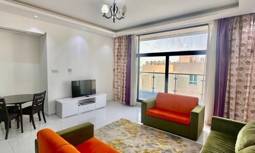 A modern living room with white walls, a patterned rug, orange and green sofas, a TV on a white stand, a chandelier, and sliding doors with curtains leading to a balcony.