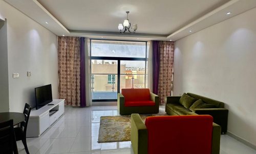 A modern living room with tiled floors, a green and red sofa set, a TV on a white stand, a dining table with chairs, and large windows with brown curtains.