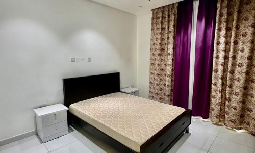 A minimalist bedroom with a bed frame and mattress, flanked by two white nightstands. The room features tiled flooring, a chandelier, and a window with purple and patterned curtains.