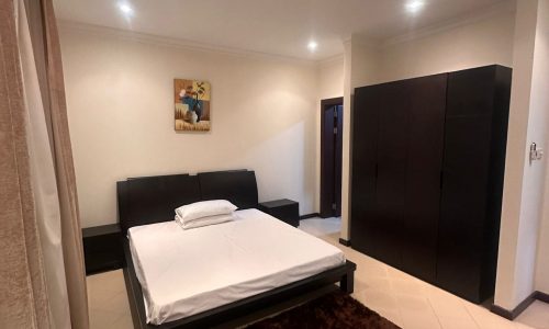 A minimalist bedroom with white walls, a large bed with white bedding, a dark wooden wardrobe, nightstands, brown rug, and a painting above the bed. Recessed lighting illuminates the room.